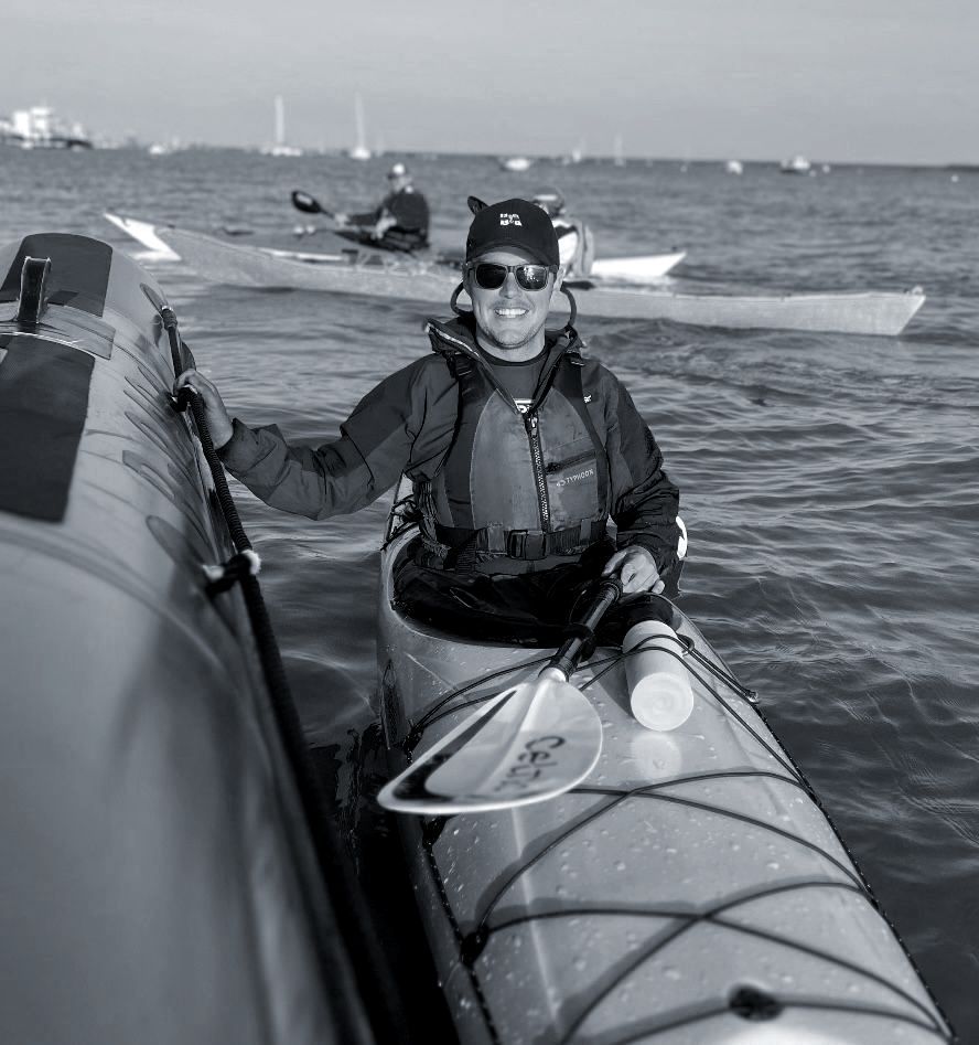 Person in kayak, wearing sunglasses and life vest, holding a paddle, near a boat on the water. Another kayaker in the background.