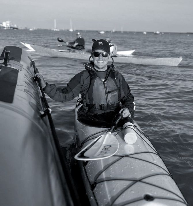 Person in kayak smiling, near boat, paddling on water, other kayakers in background.