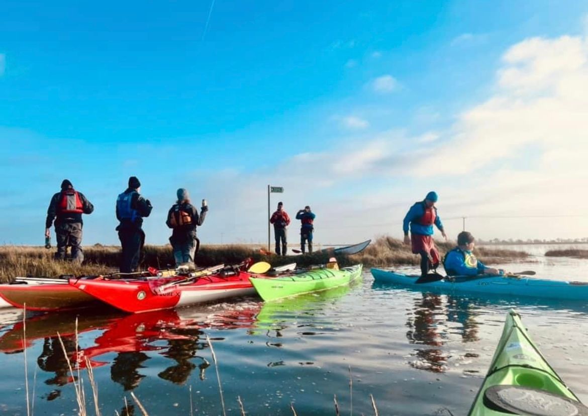 Kayakers on the water near a grassy shore under a blue sky, some standing.