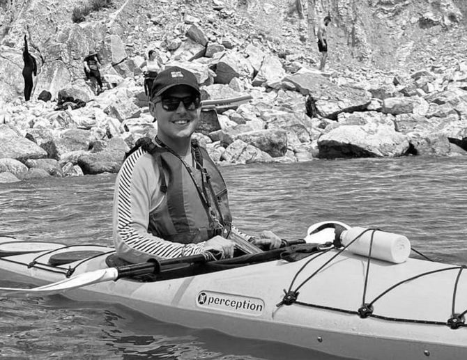 Man in kayak, smiling. Wearing sunglasses, life vest, hat. On water, rocky background.