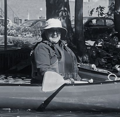 Person in a canoe paddling, wearing a hat and life vest, outdoors.