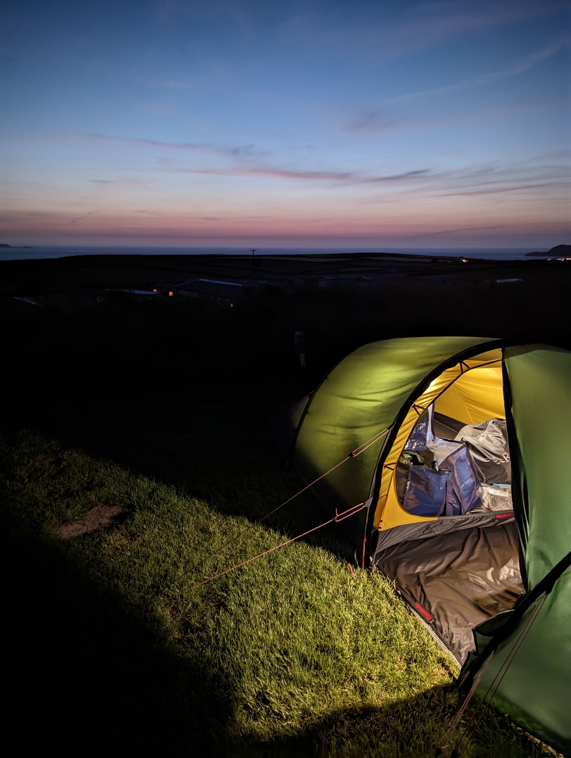 A green tent glows at dusk, open to a dark landscape and a colorful sunset.