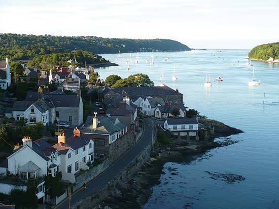 Coastal village with white buildings, road, and boats in the water; trees on opposite shore.