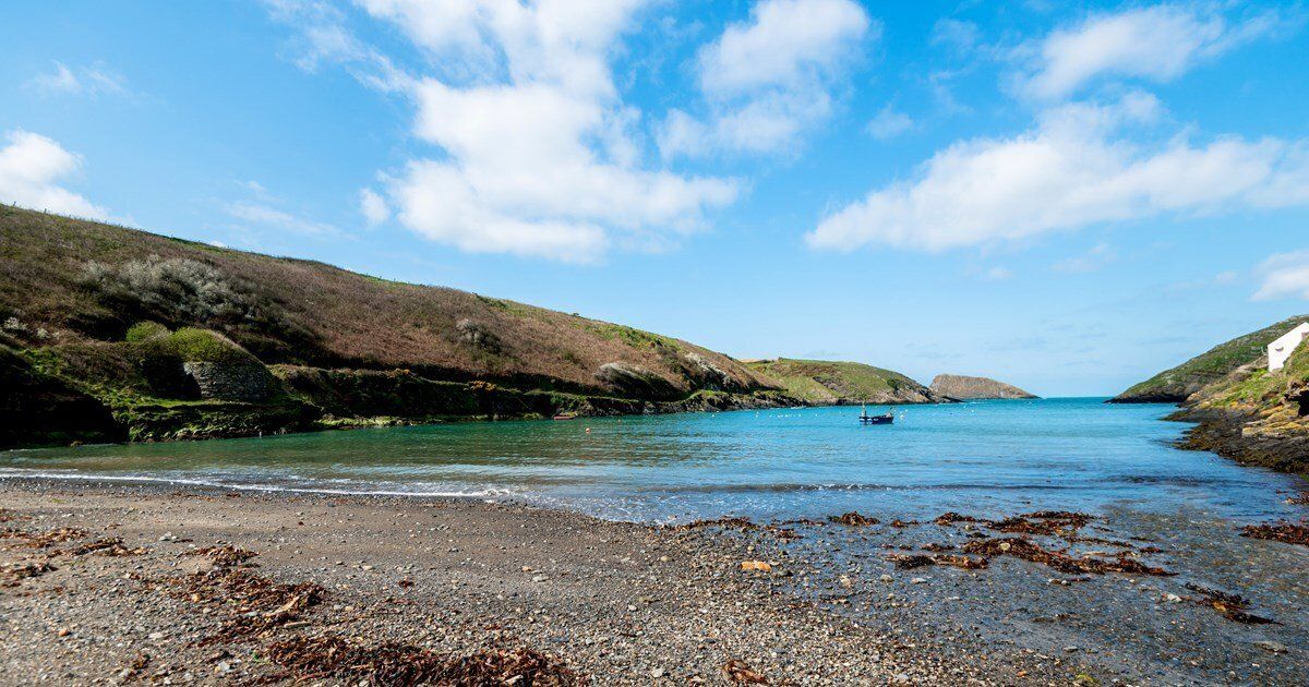 Coastal cove with pebble beach, calm blue water, hills, and a partly cloudy sky.