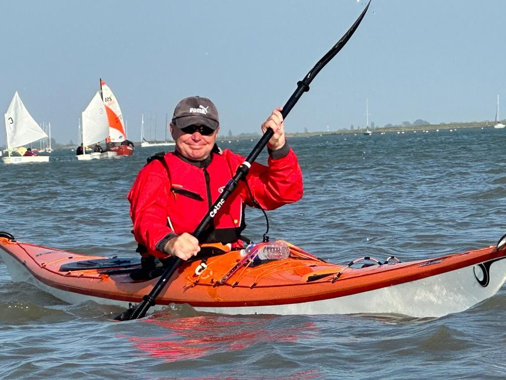 Man kayaking in orange kayak, wearing red jacket, paddles on water, sailboats in background.