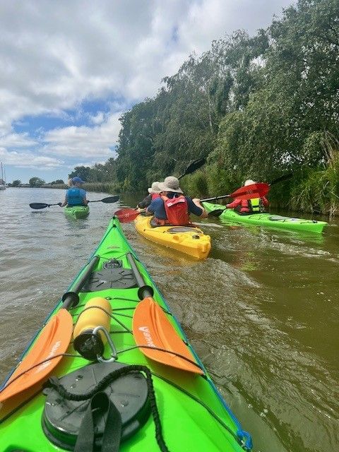 Kayakers paddle on a river with lush green banks under a cloudy sky.