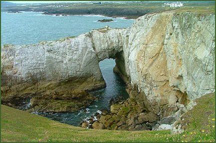 Natural arch in white rock over blue water. Green grass in foreground, other land in the distance.