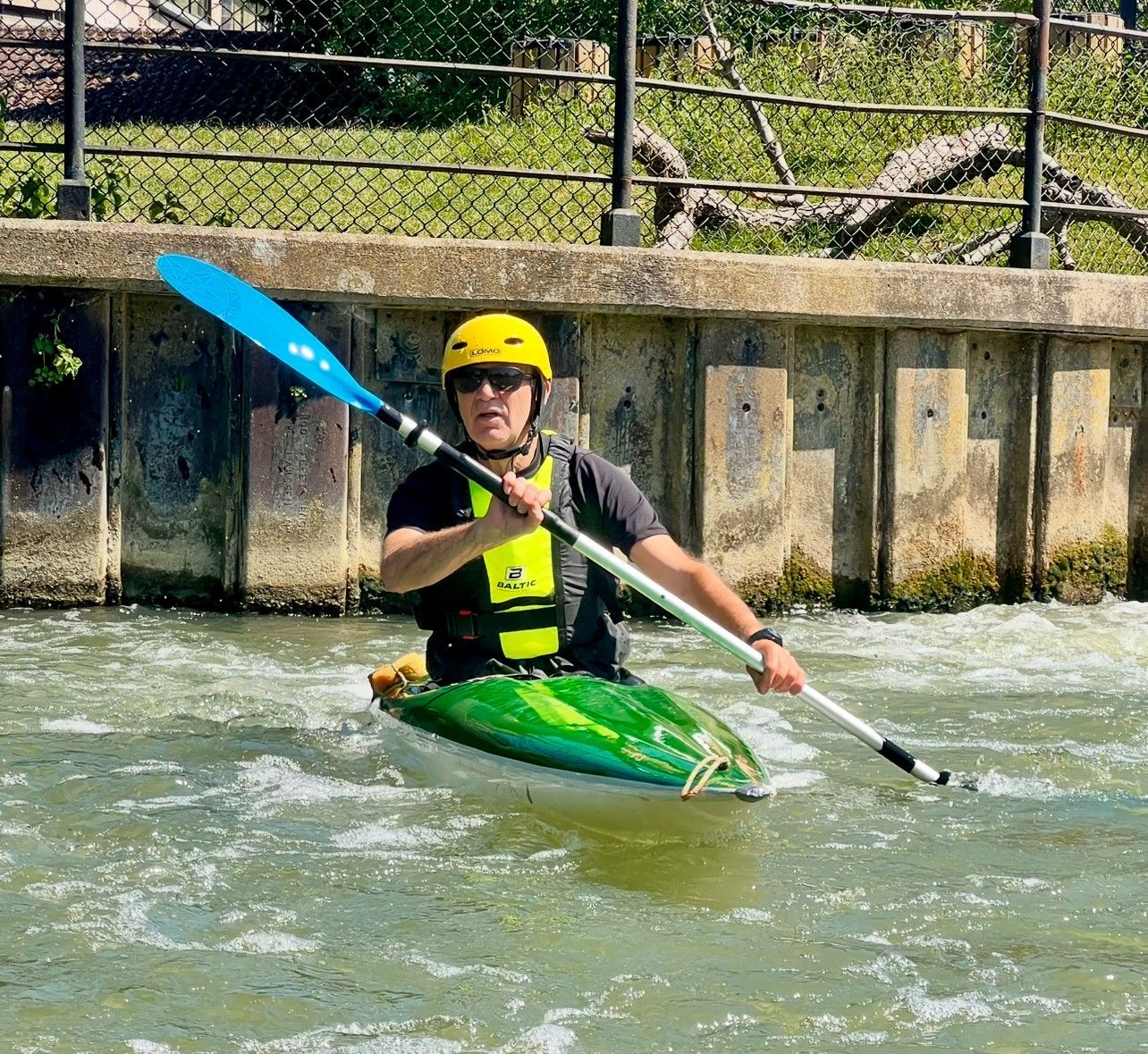 Person in a kayak with blue paddle, wearing a helmet and life vest, paddling in a water channel.