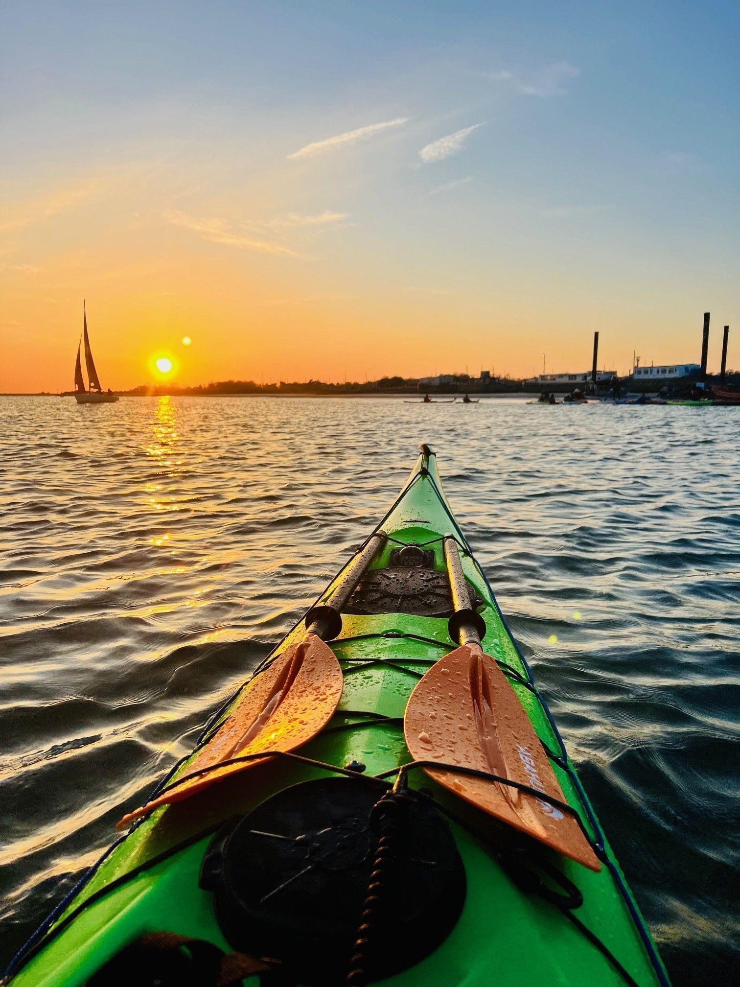 Green kayak on water at sunset, paddles resting. Sun near horizon, buildings in the distance.