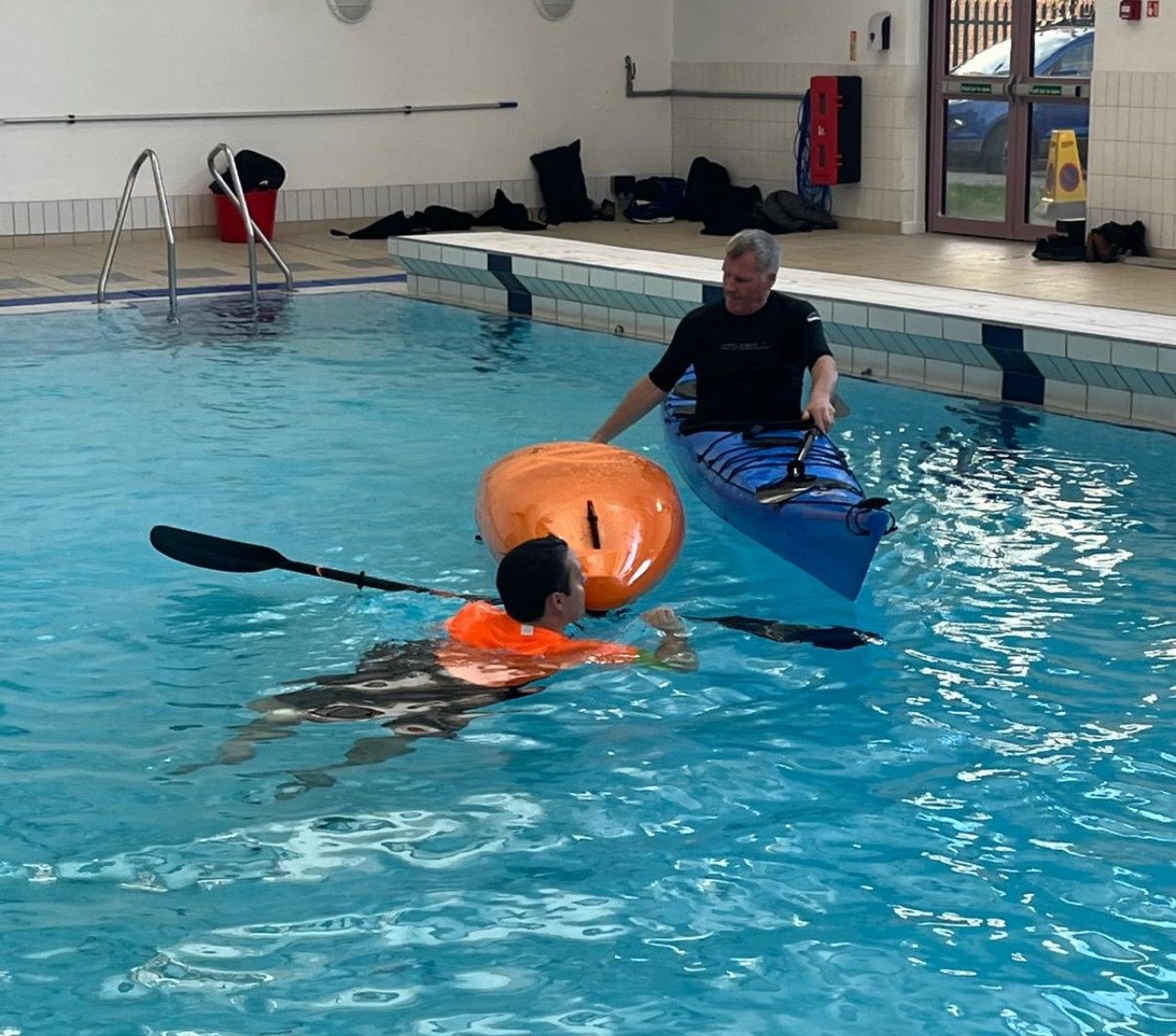 A man in a kayak instructs another in the pool. An orange float nearby.