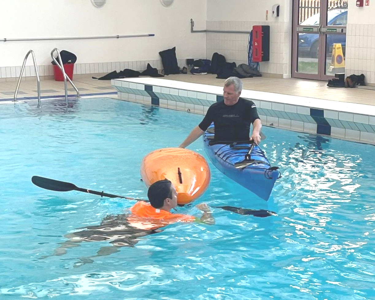 Man in kayak assisting another person in pool with orange float and paddle.