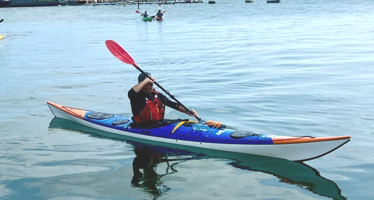 Person kayaking on calm water with a red paddle. Other kayakers and docks visible.