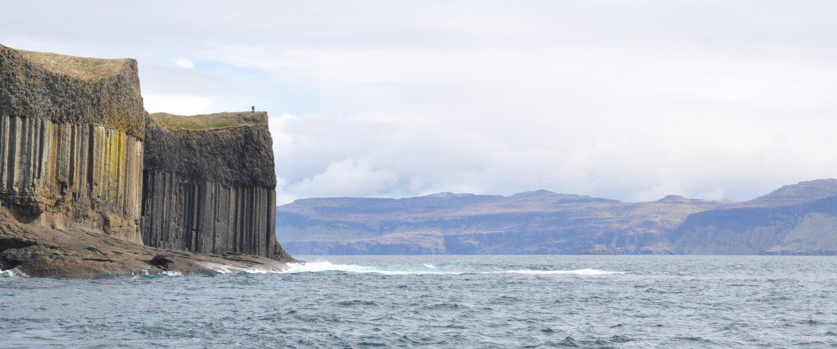 Coastal basalt columns, person on top. Cloudy day, waves in sea, mountains in background.