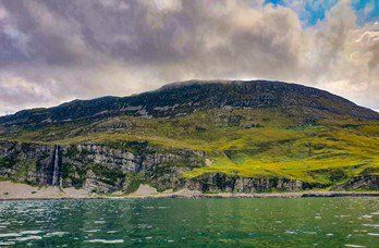 A mountain with a waterfall, grassy slopes, and cloudy sky over a body of water.