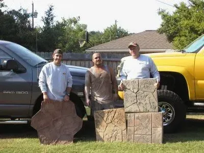 Three men holding concrete pavers, posing in front of two trucks, on a grassy lawn.