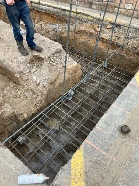 Construction site: Man standing near an excavated trench with steel rebar framework. Dark gray metal is laid horizontally and vertically, secured with gray plastic spacers.
