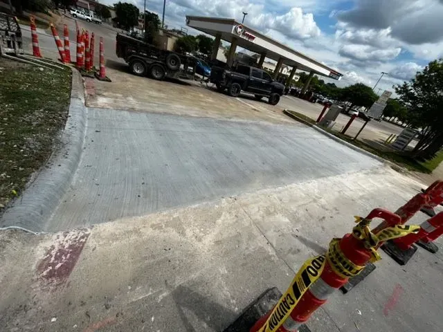 Concrete work in progress at a gas station. A truck and trailer are parked nearby. Orange cones and caution tape are present.