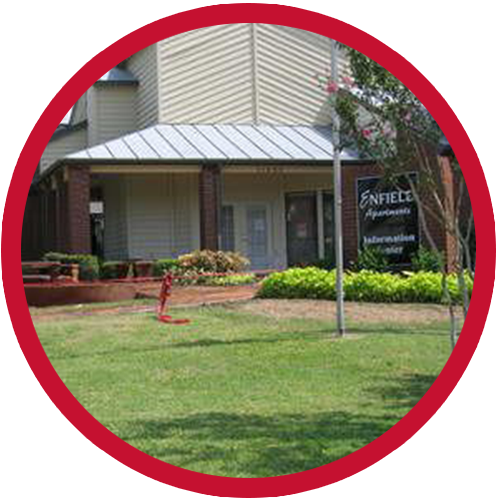 Exterior of Pine Hills Apartments office building, light beige with brick accents, surrounded by green lawn and landscaping.