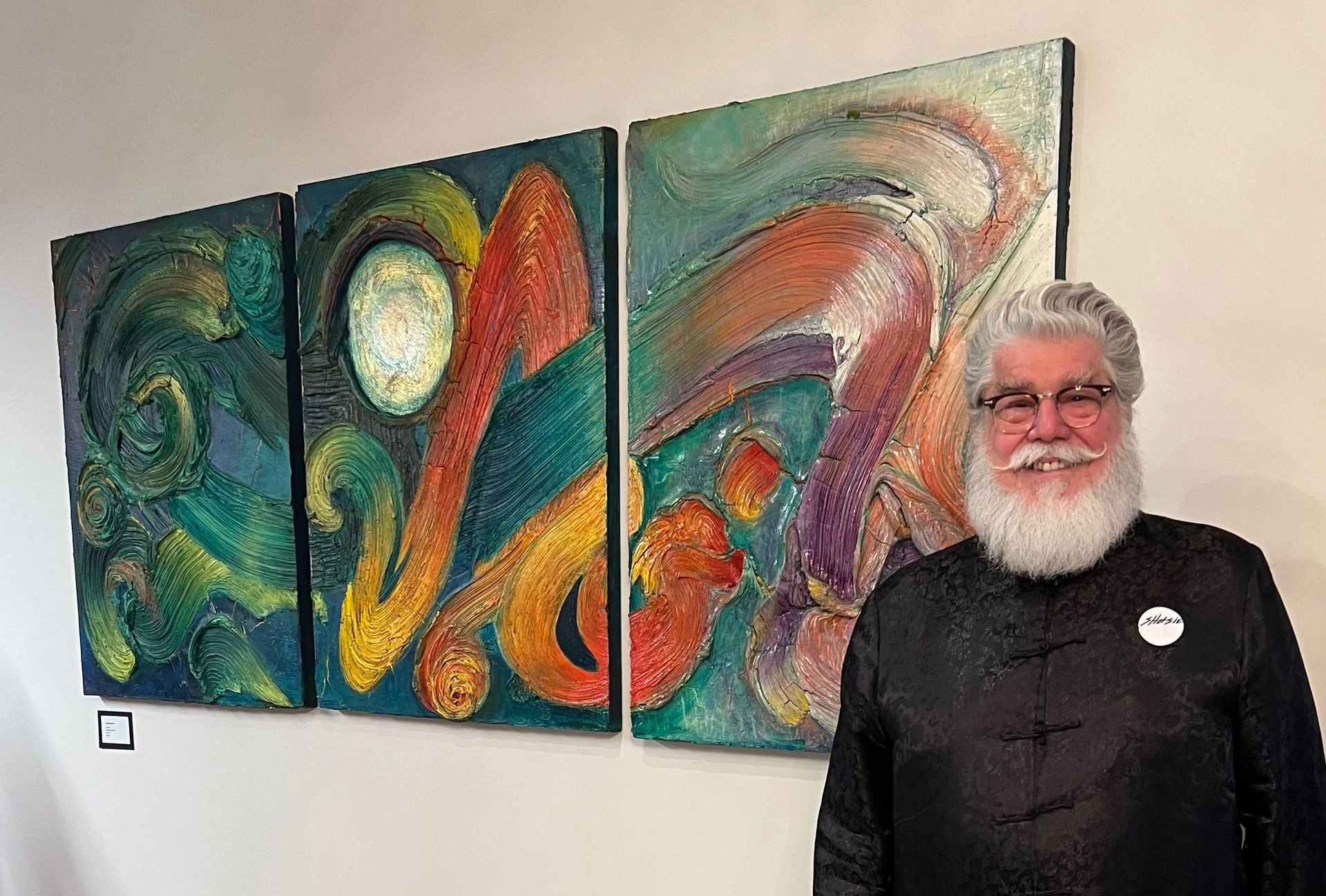 A man with a beard is standing in front of three paintings on a wall.