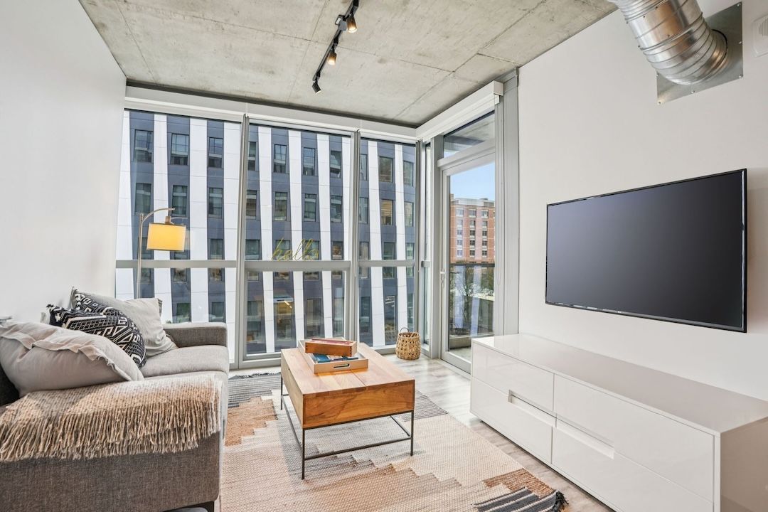 Modern living room with large windows, gray sofa, white TV console, and wooden coffee table