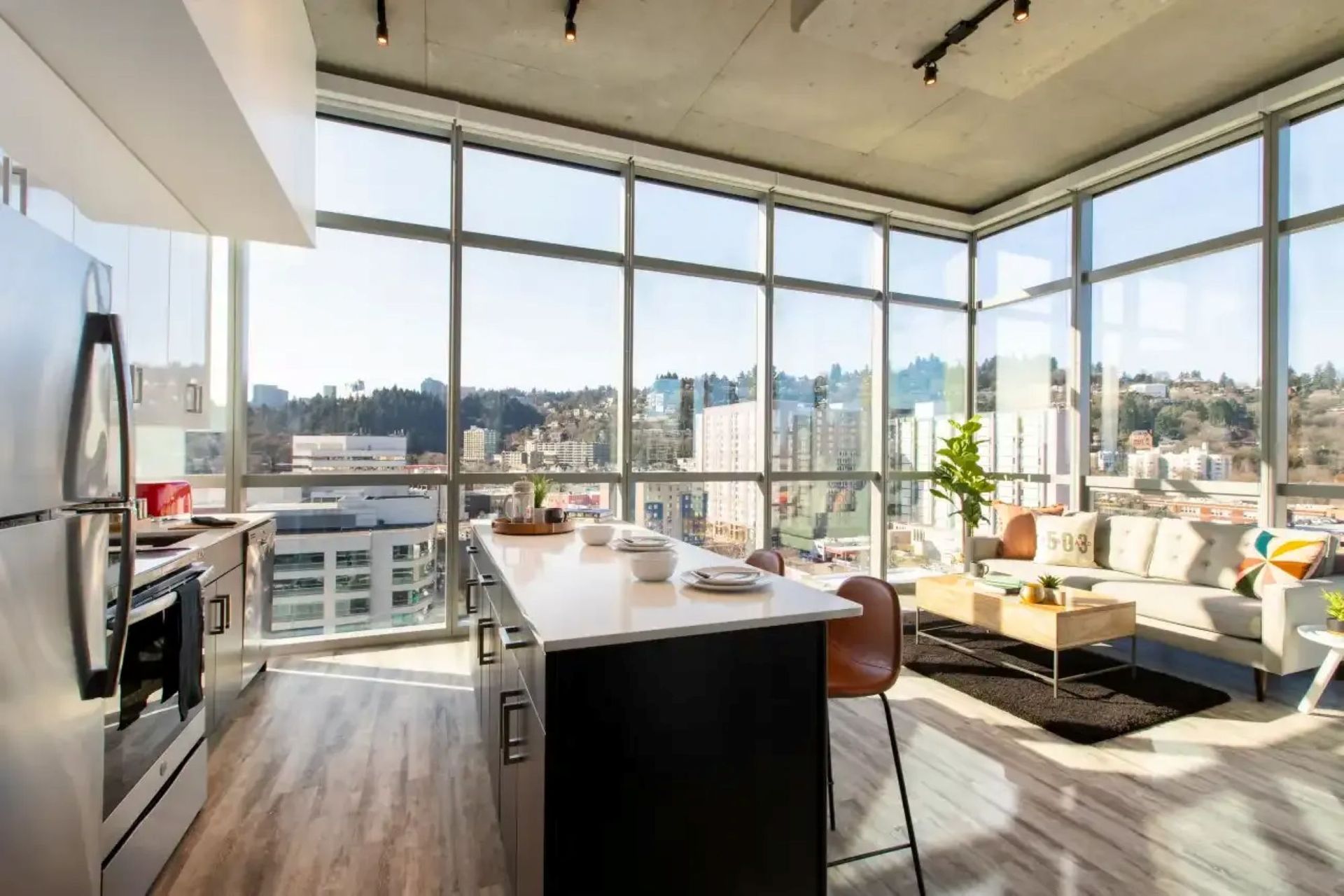A sunlit apartment featuring floor-to-ceiling windows, a modern kitchen island, and scenic city sky