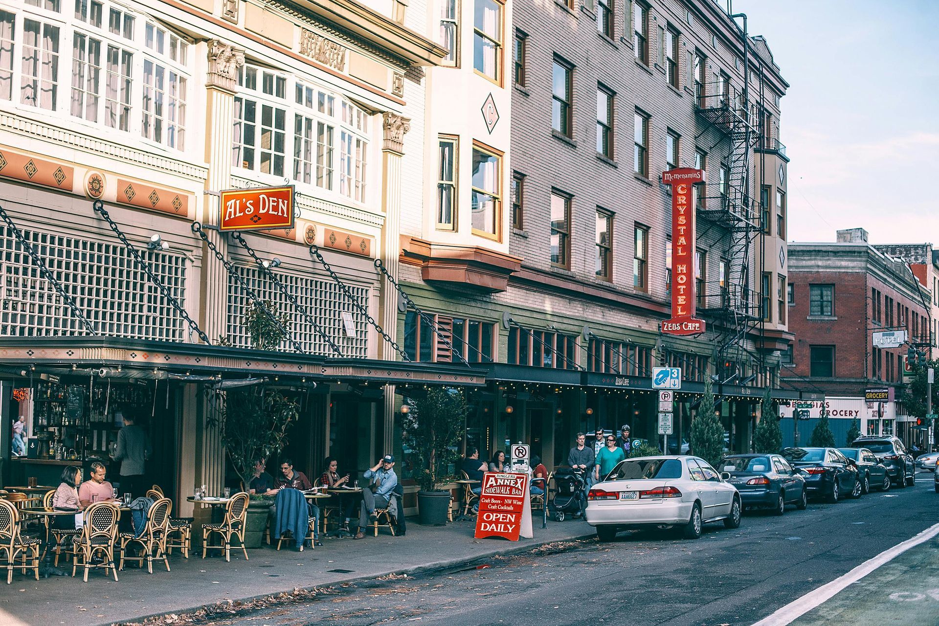 People sitting at tables outside a bustling sidewalk cafe alongside parked cars and historic city.