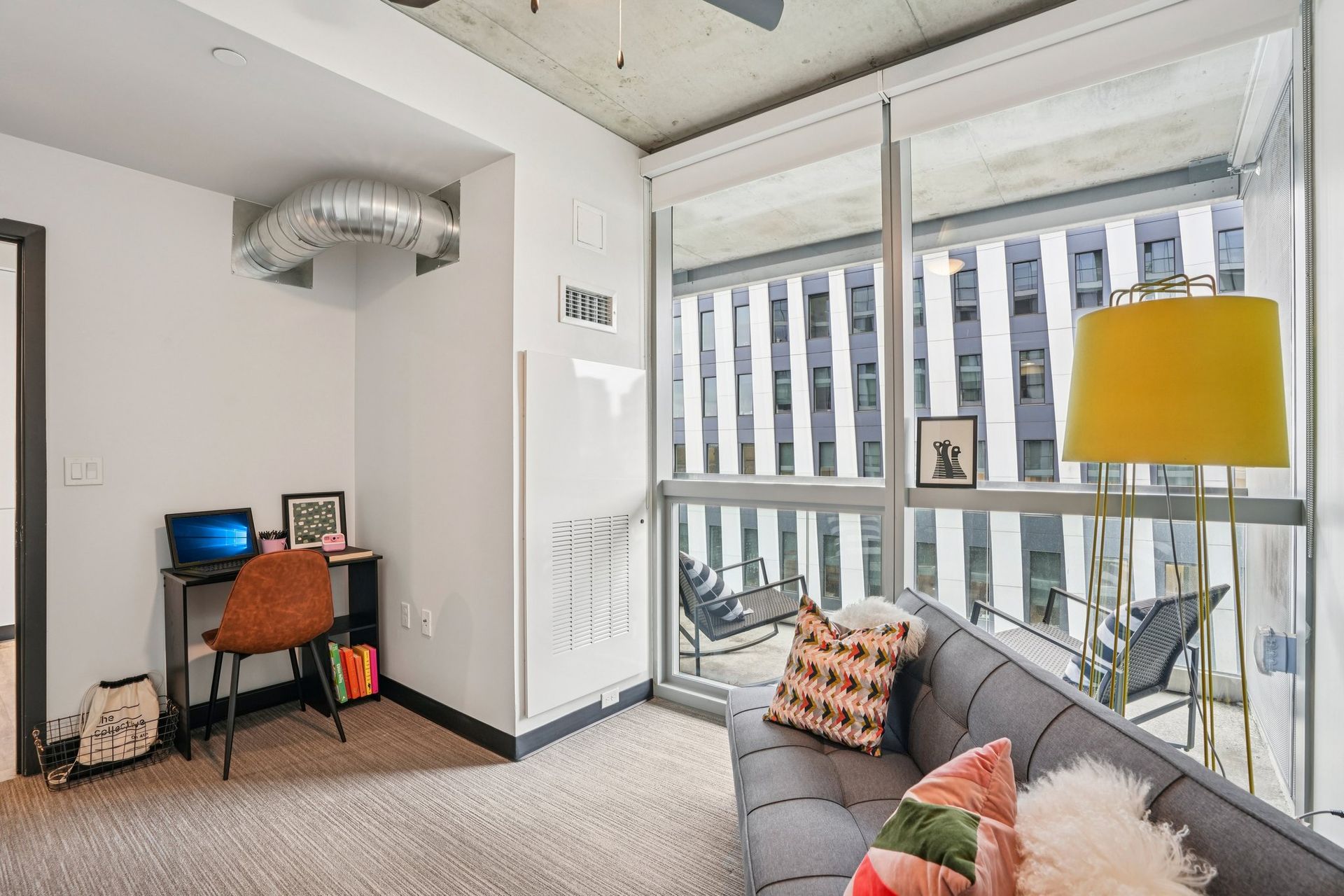 Small office corner beside a bright window, with desk, orange chair, gray sofa, and yellow lamp.