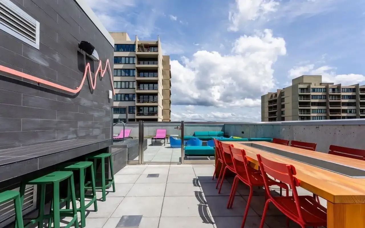 A rooftop terrace featuring colorful outdoor chairs, a long table with ample seating and a clear blue sky from a high-rise apartment.