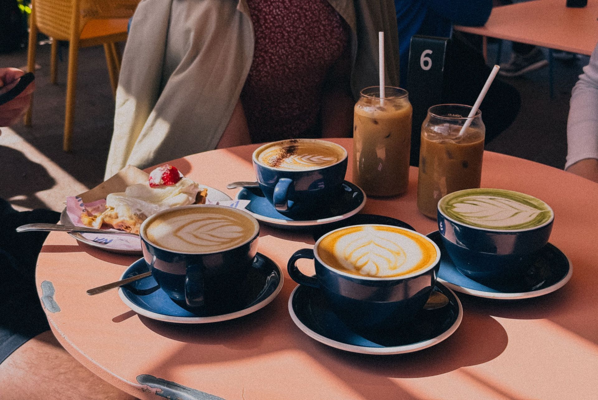 Four blue cups of coffee featuring latte art sitting on a pink table near iced drinks and a strawberry pastry.