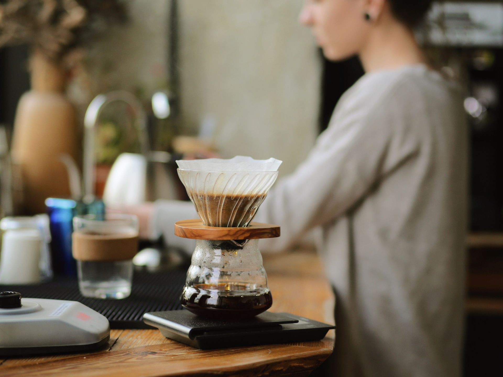 A person preparing fresh pour-over coffee using a glass dripper and carafe resting on a digital scale in a cafe.