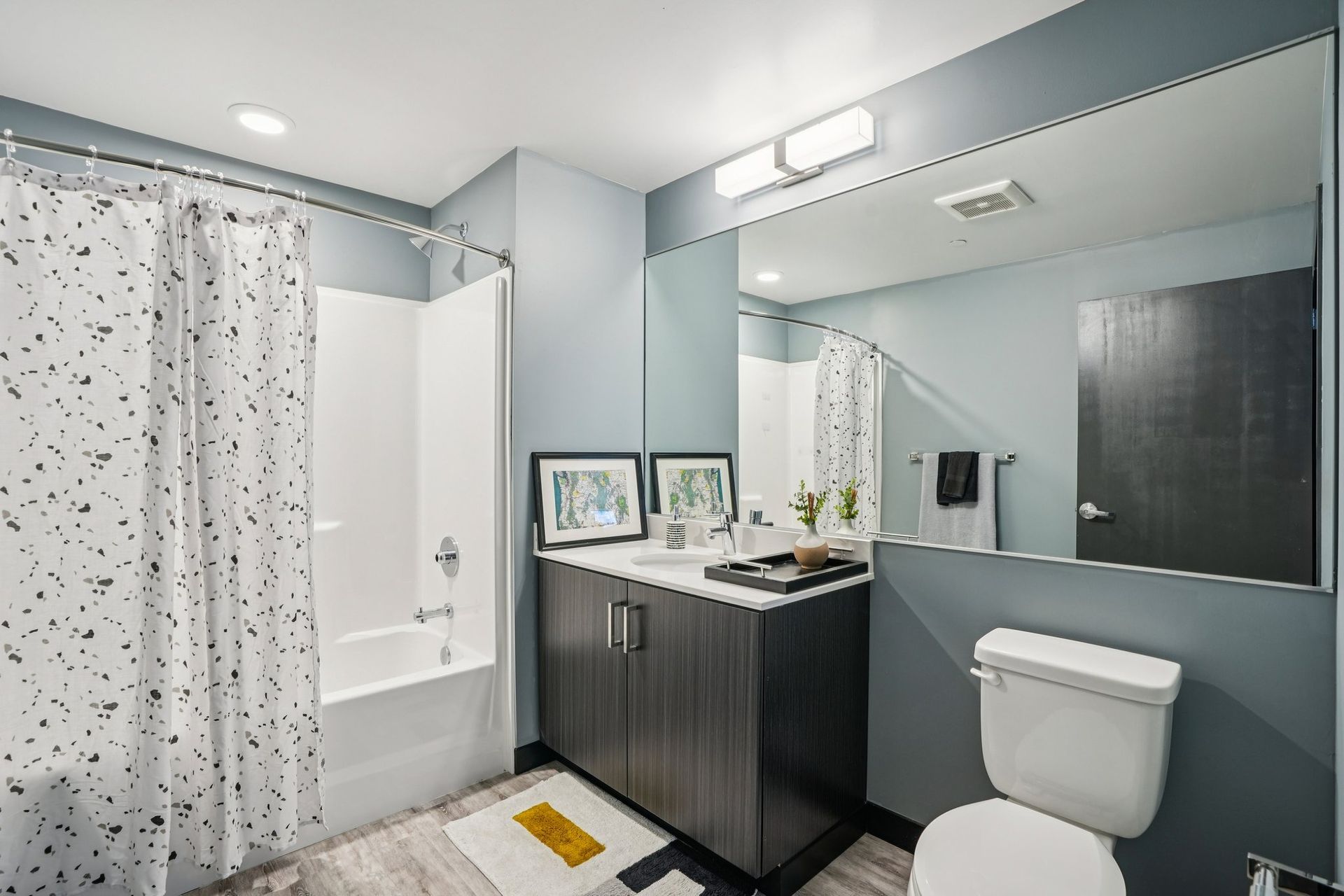 Modern bathroom with dark vanity, large mirror, white tub, and toilet on gray walls.