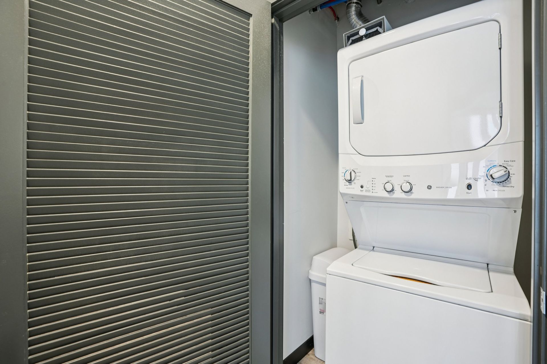 Compact apartment kitchen and entryway with wood floors, white cabinets, gray door, and a small adjacent room.