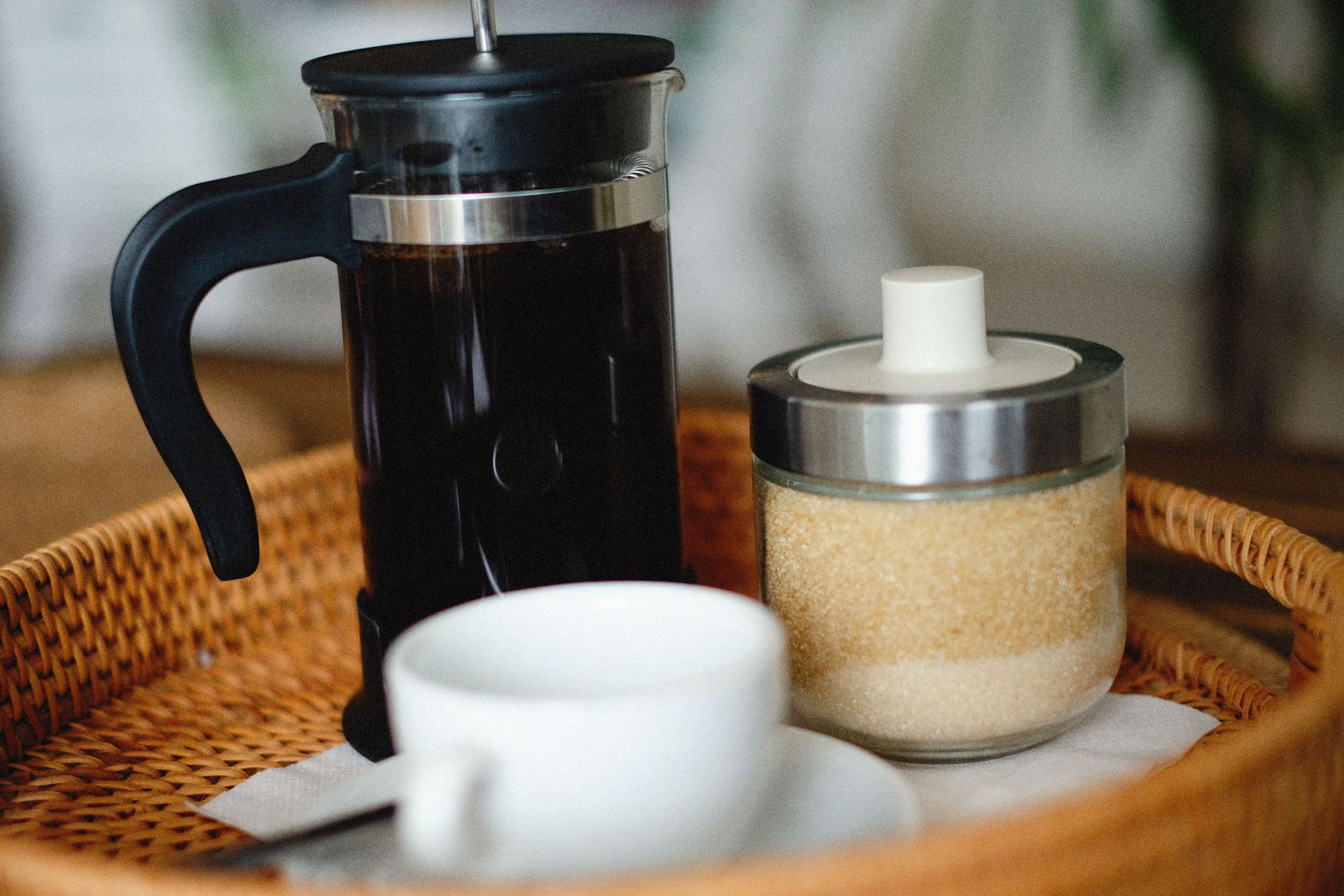A French press filled with dark coffee sitting beside a white cup and sugar container inside a woven tray.