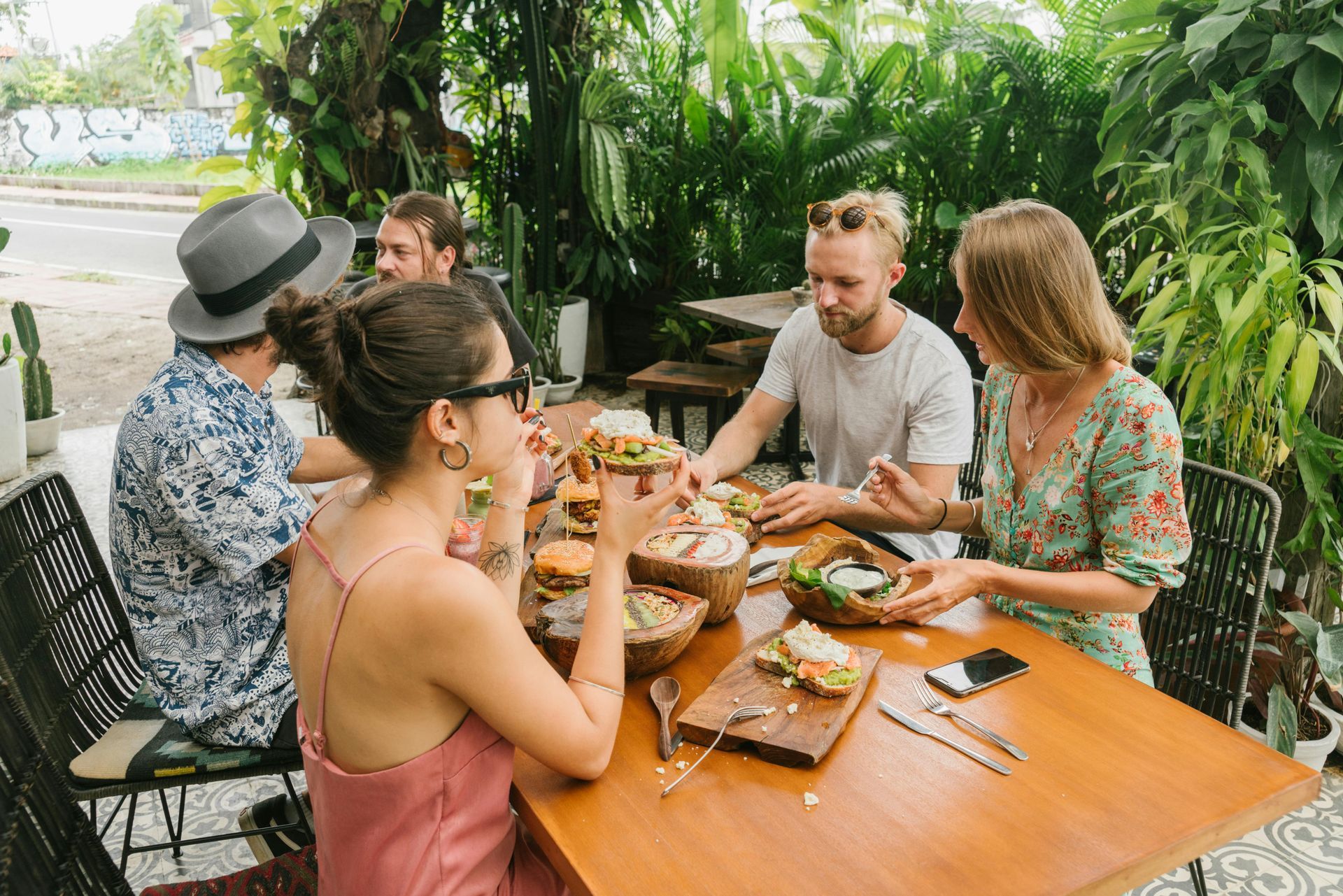 A group of friends eating healthy meals and talking while sitting around a wooden table on a lush green patio.