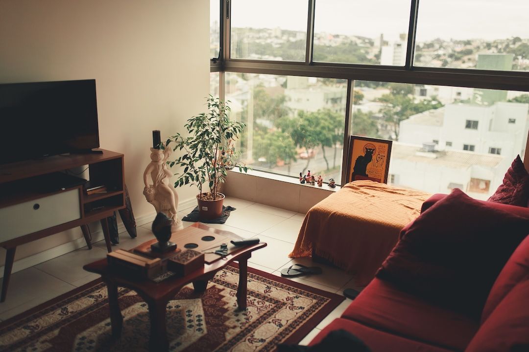 A cozy living room showing a red sofa and a wooden coffee table next to a large window overlooking a city landscape.