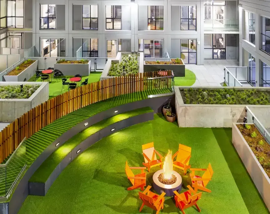 Rooftop courtyard with green turf, fire pit, orange chairs, and wooden fence. Apartment windows in the background.