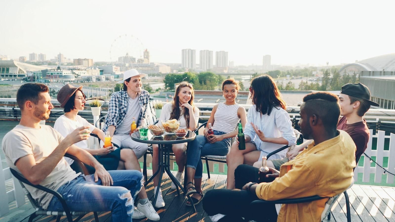 Friends chatting at an outdoor café with drinks, overlooking a city skyline on a sunny day