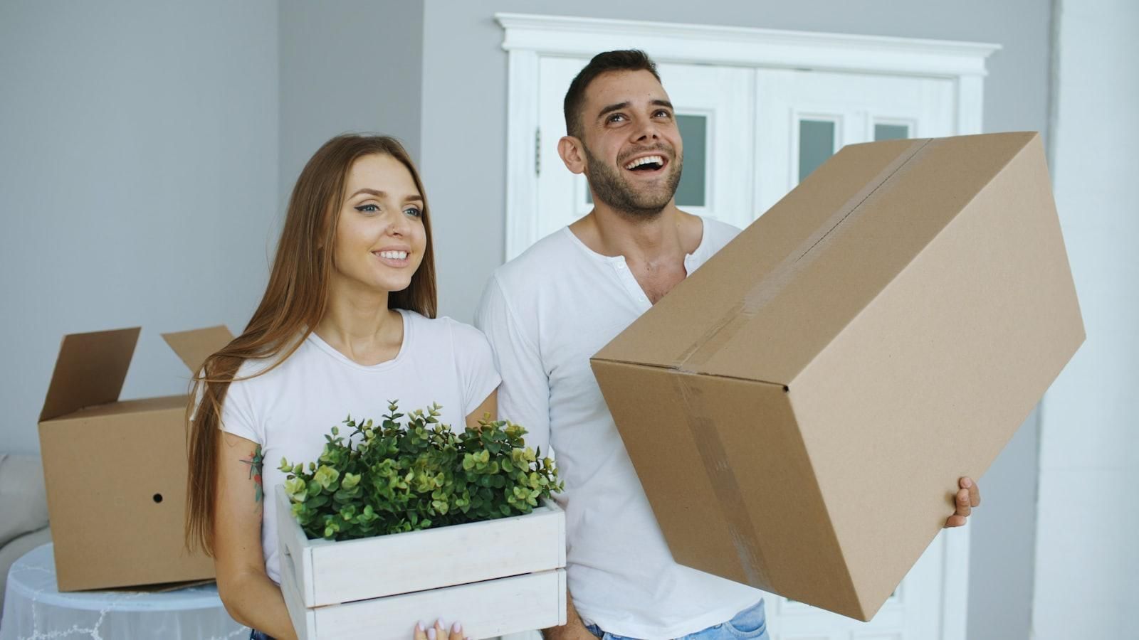 Young couple holding moving boxes and a houseplant on move-in day in their new apartment