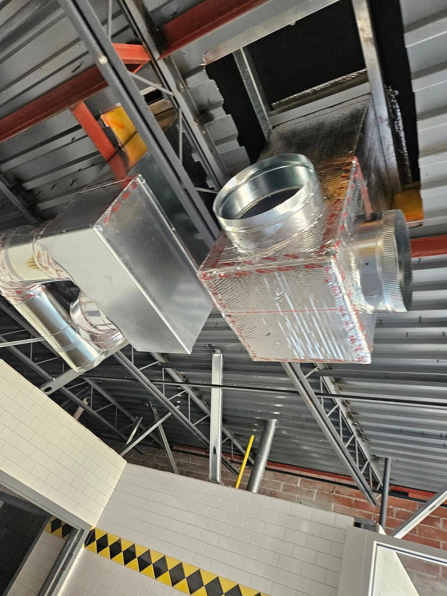 Industrial ceiling with metal ductwork, vents, and exposed beams above a hazard-striped wall.