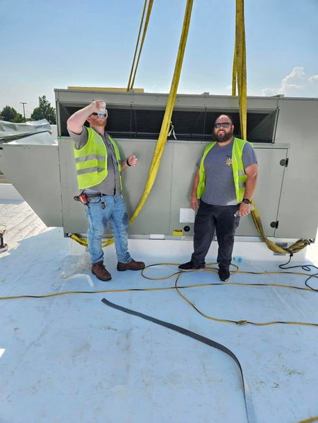 Two workers in safety vests standing on a rooftop near a large HVAC unit, with lifting straps overhead.