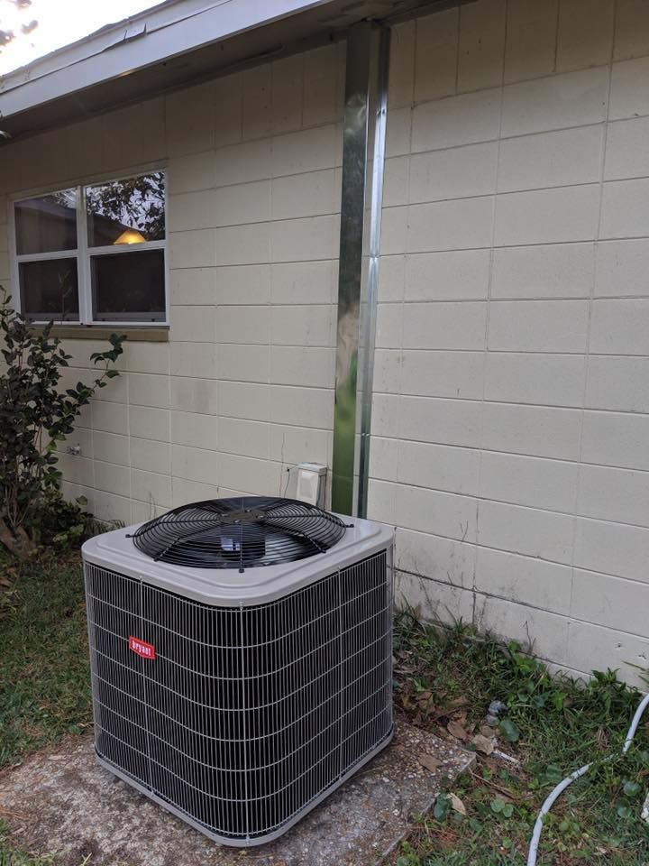 Outdoor air conditioner unit beside a beige house wall and window, with a vertical pipe behind it.