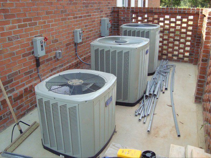 Outdoor HVAC units beside a brick wall, with electrical boxes and stacked metal pipes on a concrete pad.