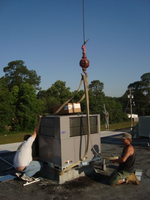 Workers rigging a large gray equipment unit with a crane outdoors under a clear sky