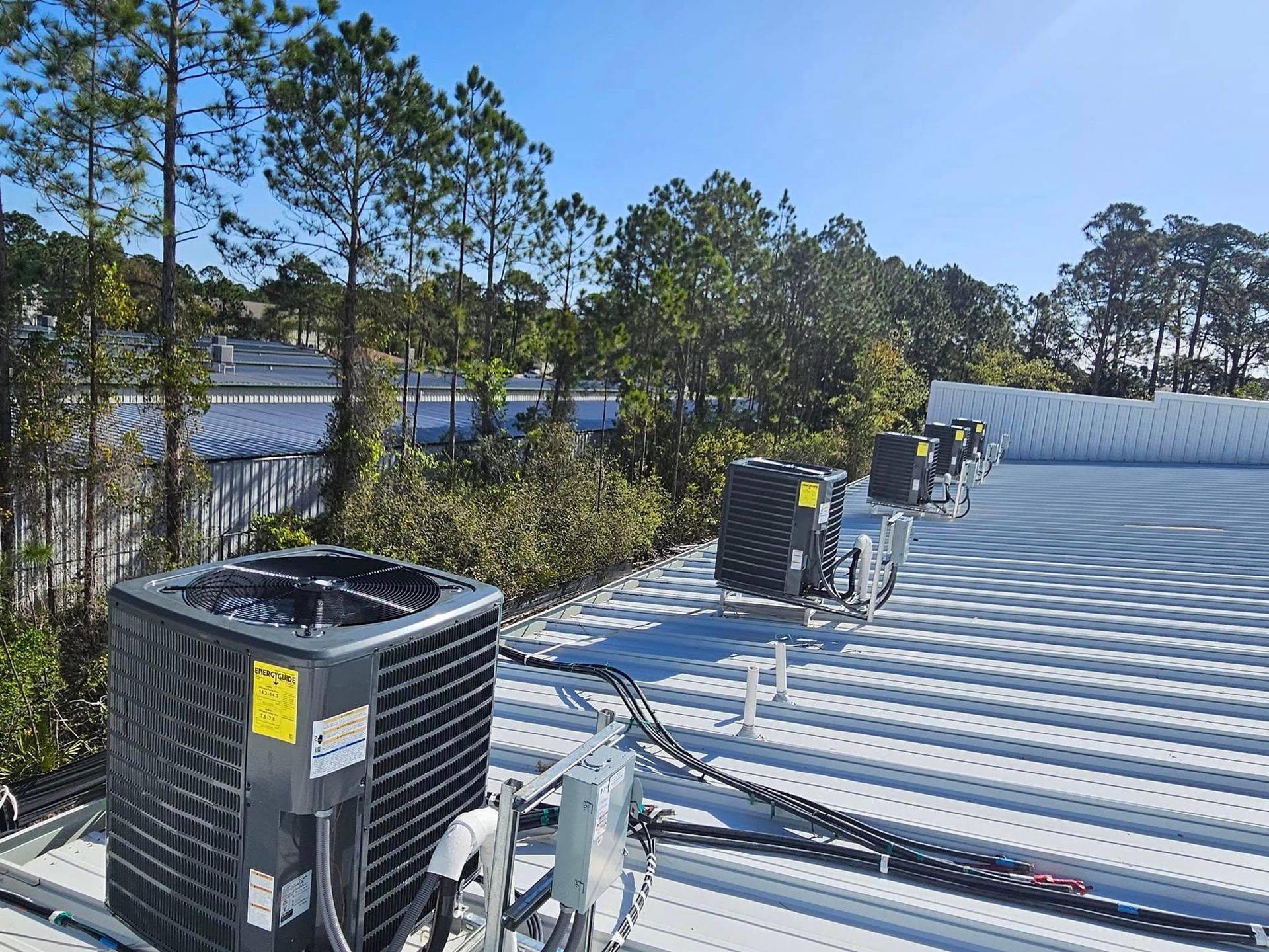 Rooftop HVAC units on a metal roof beside trees and a body of water under a clear sky