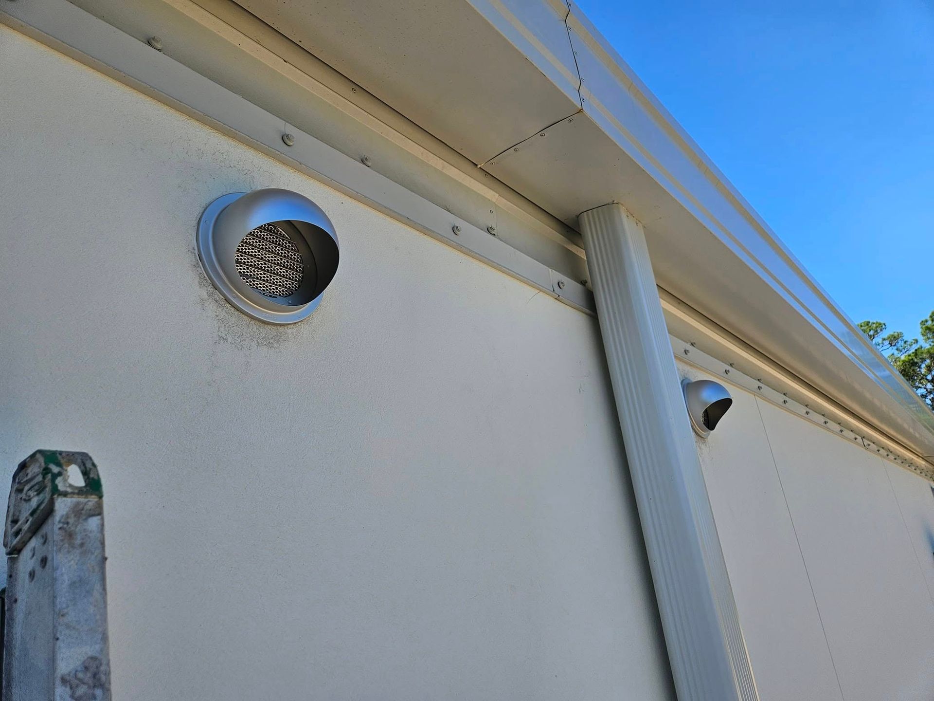 White exterior wall with a round vent and vertical downspout against a blue sky.