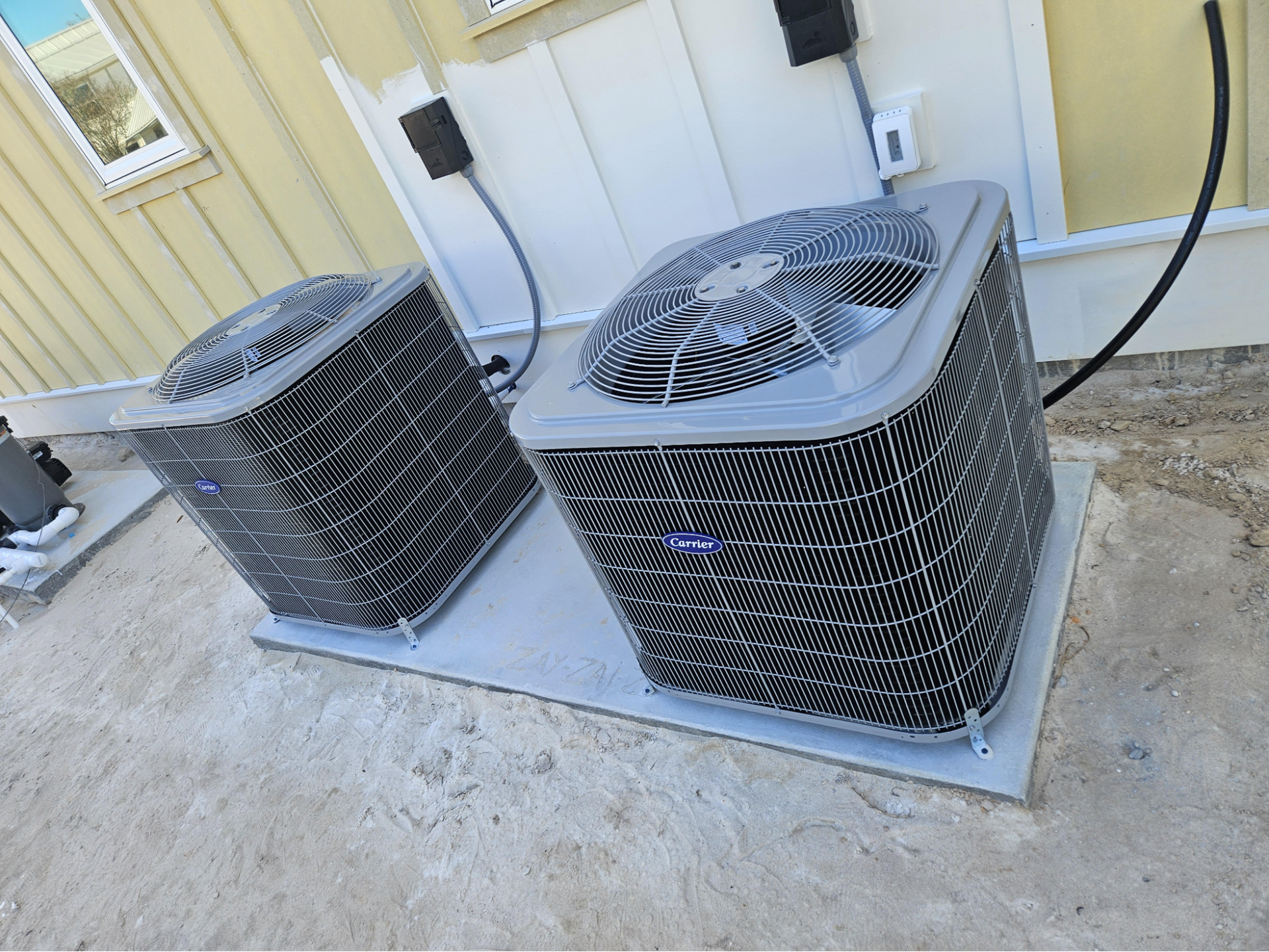 Two outdoor air conditioner units on a concrete pad beside a house wall
