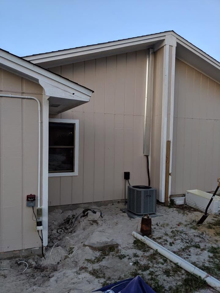 Side of a beige house with downspouts, a window, and utility equipment on a sandy patch.