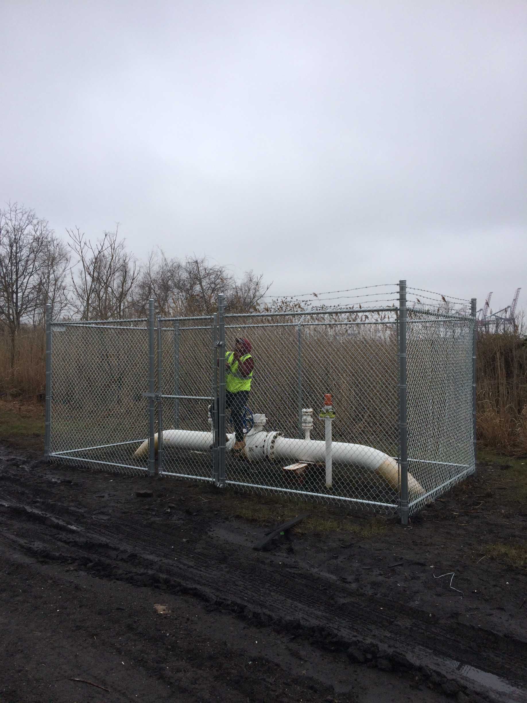Person in safety vest near fenced water pipes in muddy field. Overcast day.