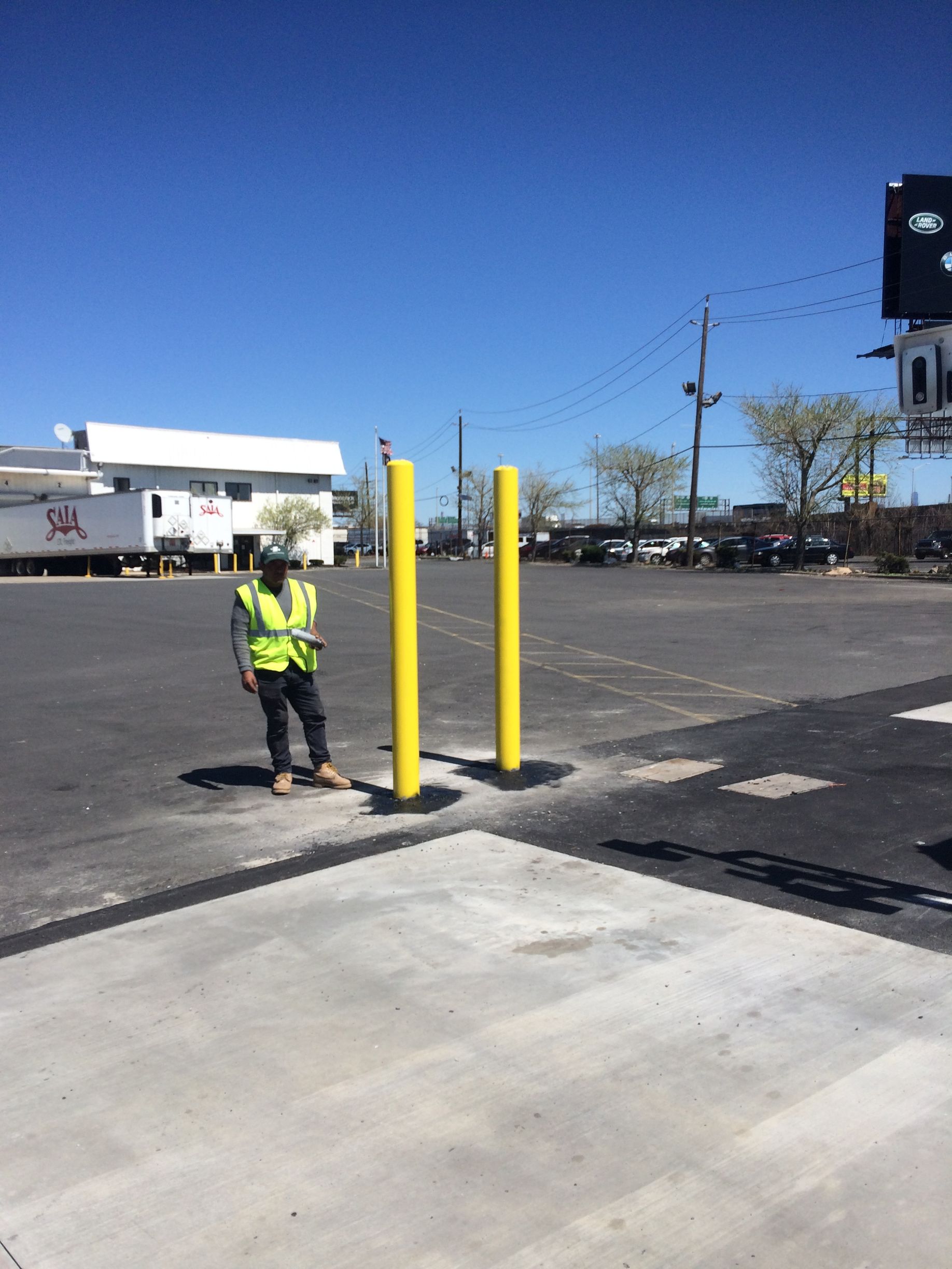 A person in a safety vest stands near two yellow posts in a parking lot.