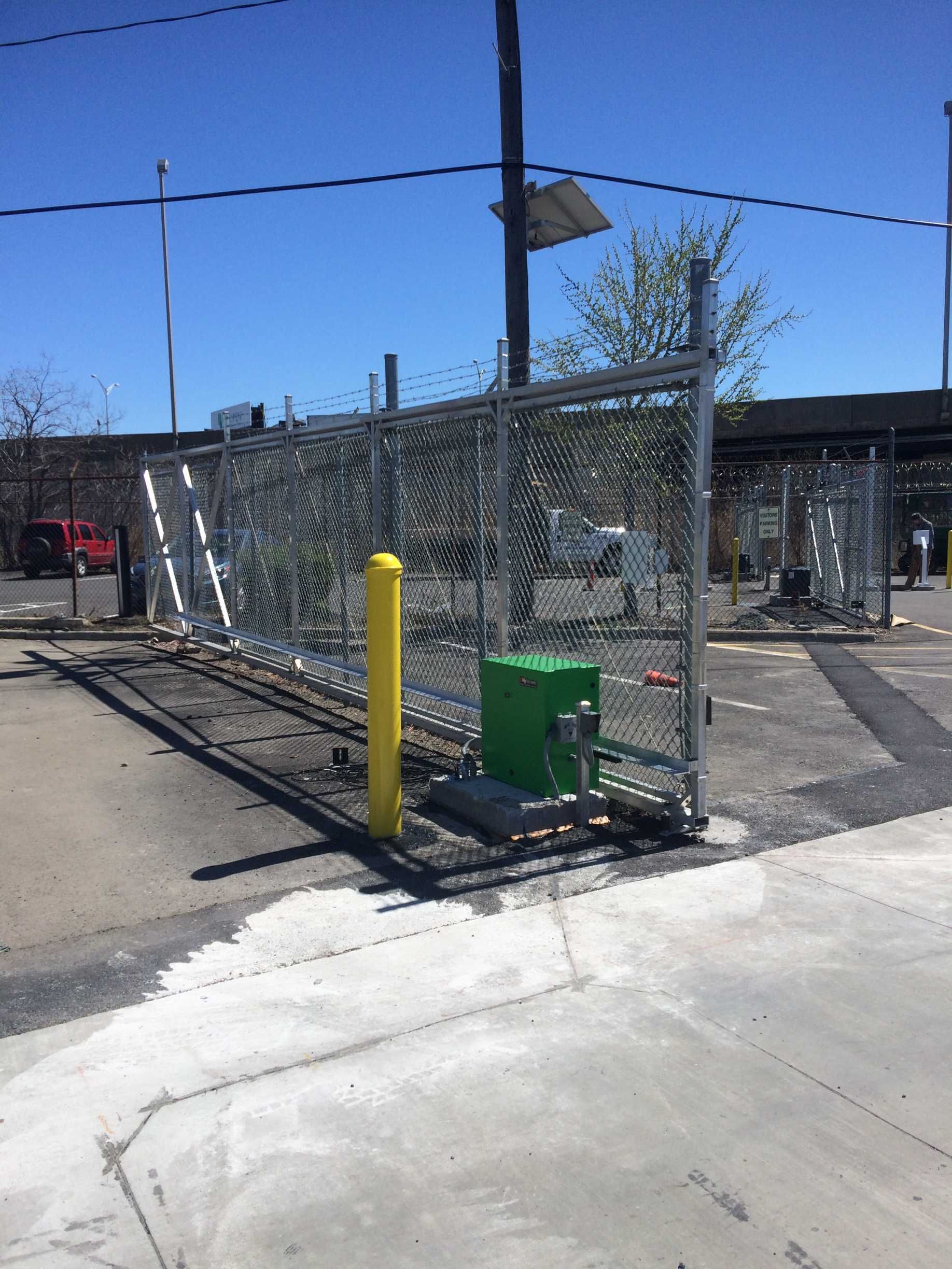 Chain-link fence with a gate, yellow bollard, and green control box in a parking lot.